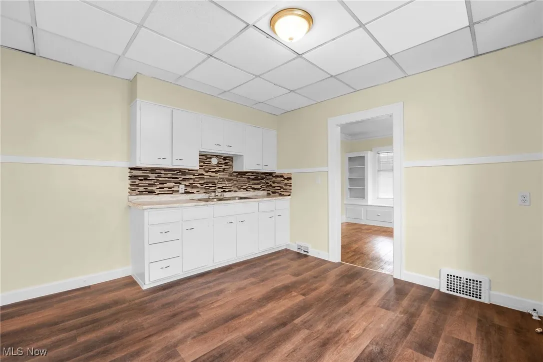 Kitchen with light countertops, dark wood-type flooring, a drop ceiling, decorative backsplash, and white cabinets