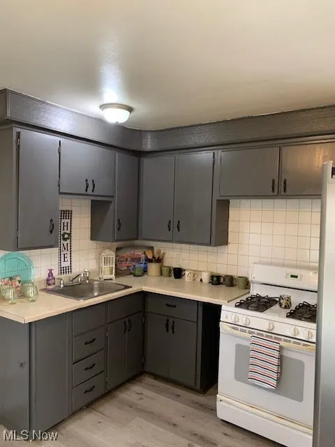Kitchen with white gas stove, decorative backsplash, light countertops, light wood finished floors, and gray cabinetry