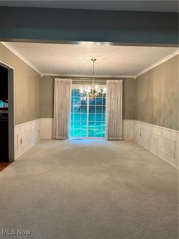 Carpeted empty room featuring crown molding, a decorative wall, a wainscoted wall, and a chandelier