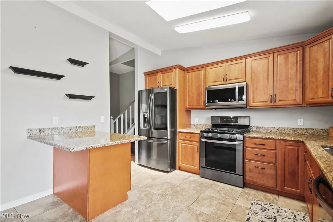 Kitchen with stainless steel appliances, vaulted ceiling, a peninsula, light stone countertops, and brown cabinets