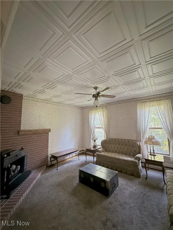 Living area with a gas  stove, an ornate tile ceiling, wallpapered walls, carpet, and a ceiling fan