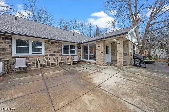 Back of house featuring a patio area, stone siding, roof with shingles, and a chimney