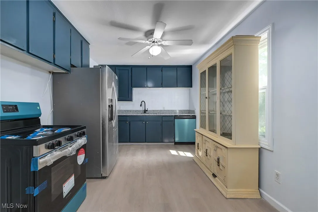 Kitchen with stainless steel appliances, blue cabinets, light wood finished floors, backsplash, and ceiling fan