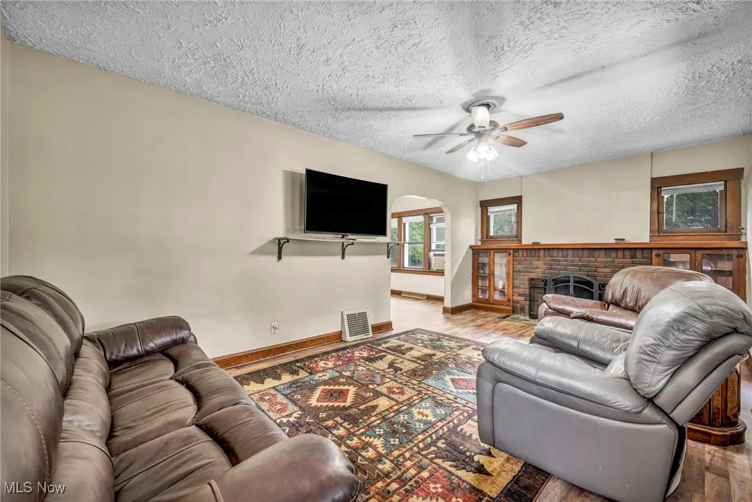 Living area with arched walkways, wood finished floors, a ceiling fan, a textured ceiling, and a brick fireplace
