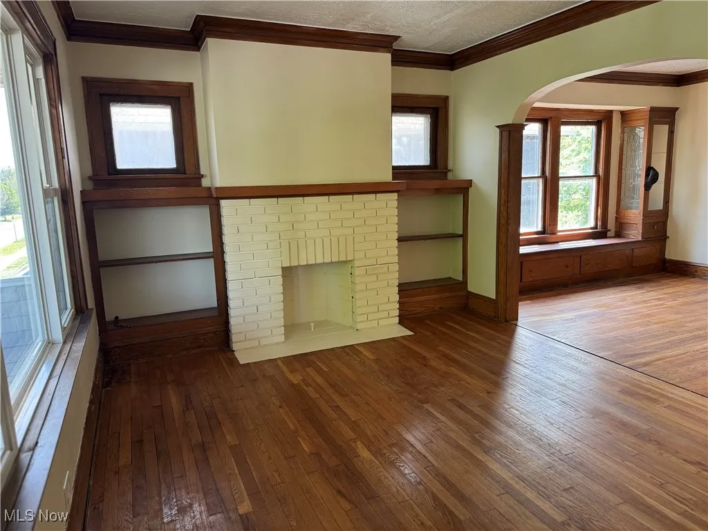 Unfurnished living room featuring hardwood / wood-style flooring, arched walkways, ornamental molding, and a brick fireplace