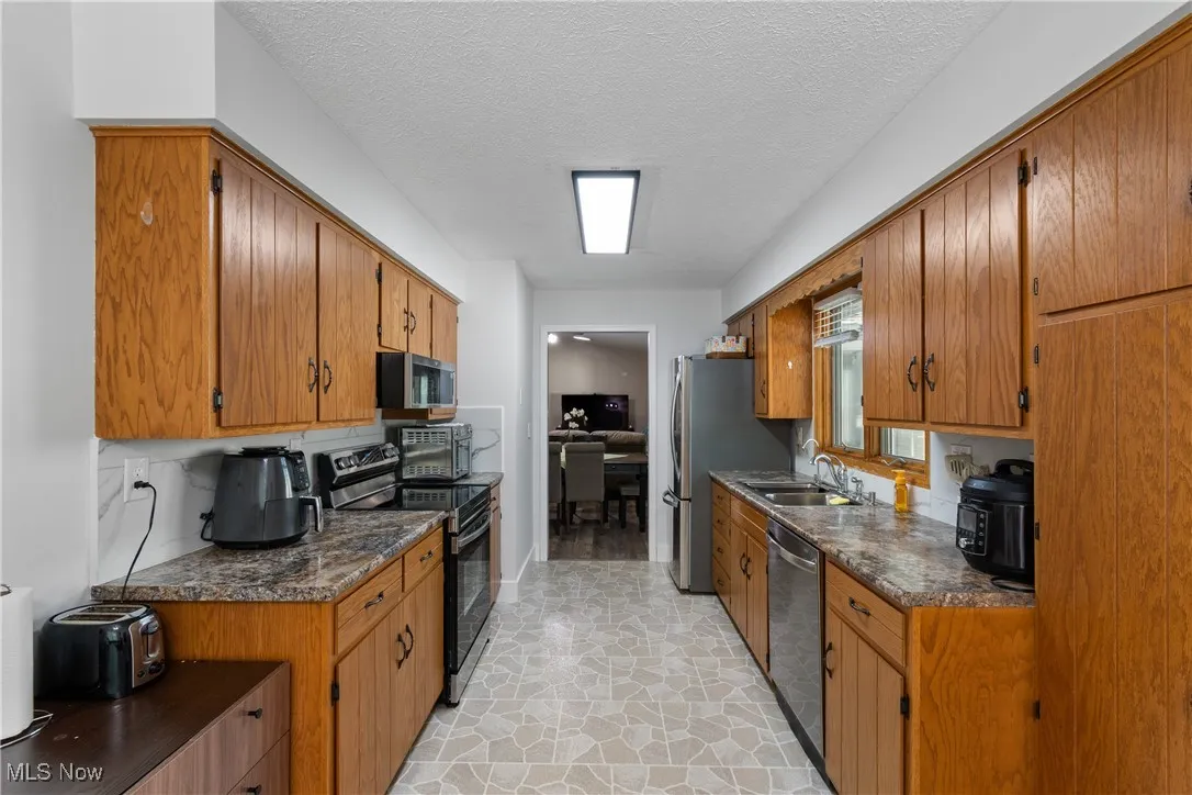 Kitchen featuring stainless steel appliances, dark countertops, brown cabinets, a textured ceiling, and light stone finish flooring
