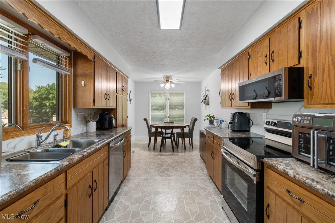 Kitchen featuring stainless steel appliances, light stone finish floors, a ceiling fan, brown cabinets, and a textured ceiling