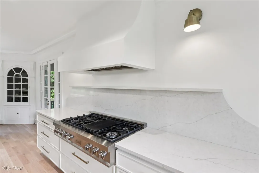 Kitchen featuring stainless steel gas cooktop, custom exhaust hood, light stone countertops, white cabinets, and ornamental molding