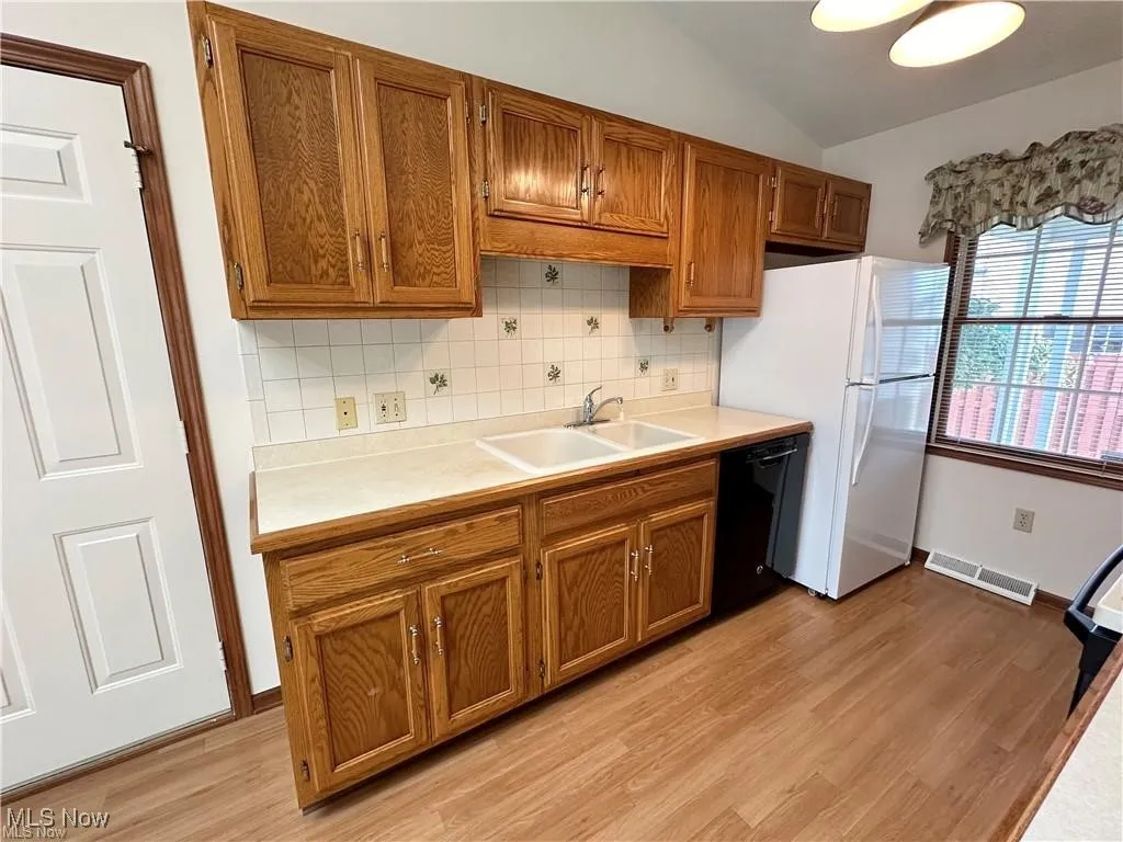 Kitchen with dishwasher, freestanding refrigerator, brown cabinetry, vaulted ceiling, and decorative backsplash