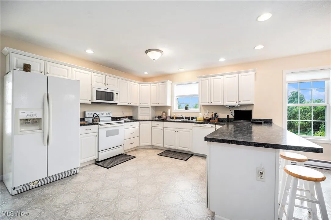 Kitchen featuring white appliances, a peninsula, dark countertops, white cabinetry, and a breakfast bar