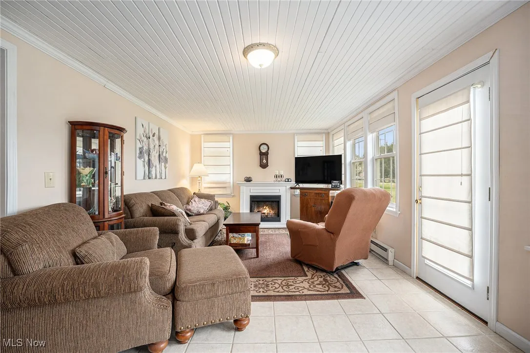Living room featuring light tile patterned flooring, a lit fireplace, ornamental molding, baseboard heating, and wooden ceiling