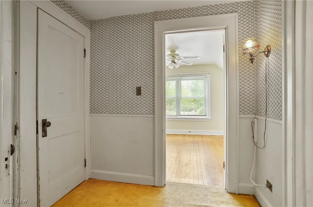 Hallway with wallpapered walls, light colored carpet, a wainscoted wall, and vaulted ceiling