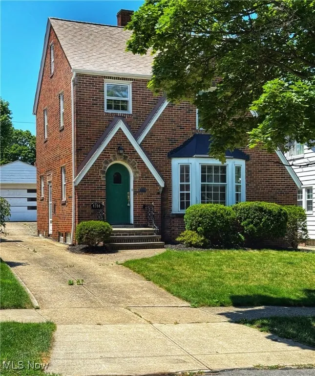 English style home featuring brick siding, a chimney, roof with shingles, a garage, and a front​​‌​​​​‌​​‌‌​‌‌​​​‌‌​‌​‌​‌​​​‌​​ lawn