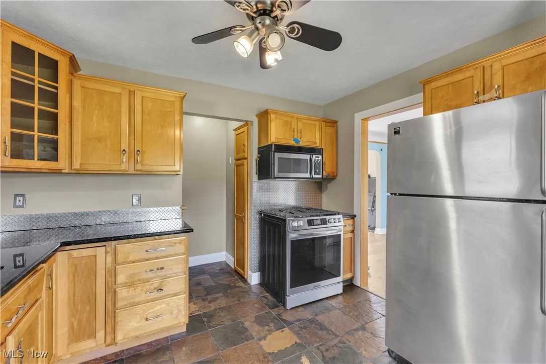 Kitchen with stainless steel appliances, ceiling fan, glass insert cabinets, stone tile floors, and decorative backsplash