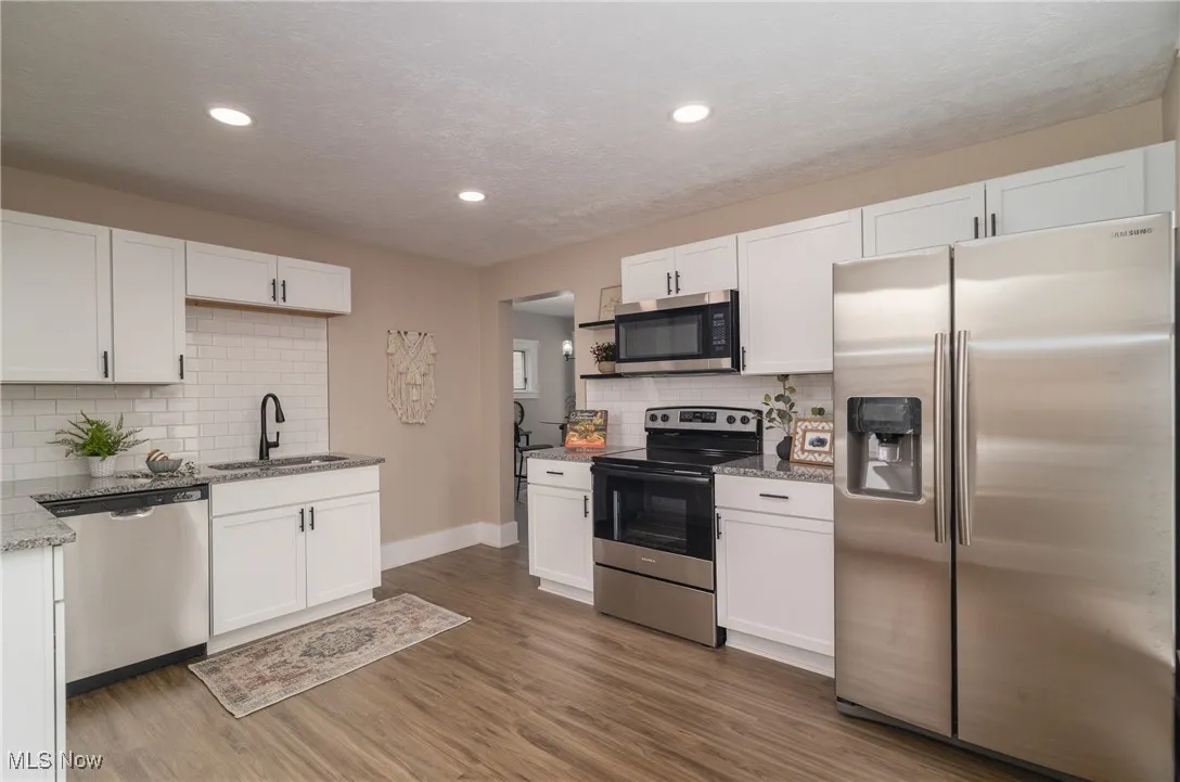 Kitchen with appliances with stainless steel finishes, white cabinetry, backsplash, light stone counters, and wood finished floors