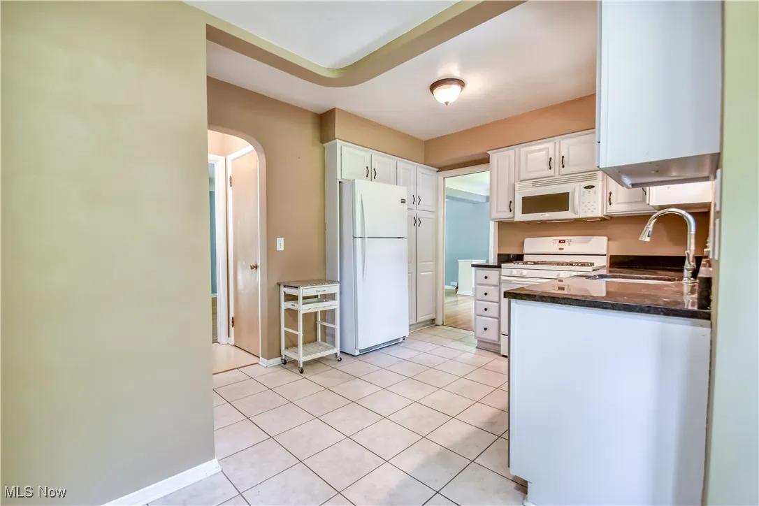 Kitchen with white appliances, arched walkways, white cabinets, light tile patterned flooring, and dark stone countertops