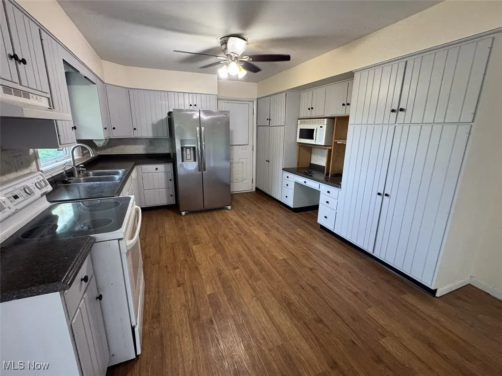 Kitchen with white appliances, dark countertops, under cabinet range hood, ceiling fan, and dark wood-style flooring