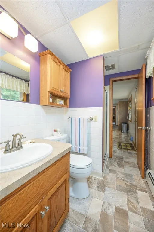 Full bath with tile walls, vanity, a stall shower, stone finish floors, and a baseboard radiator