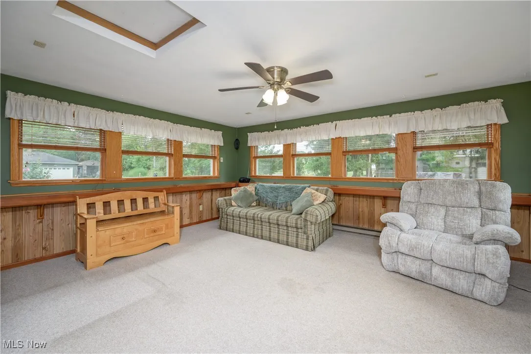 Living room featuring carpet flooring, a ceiling fan, wooden walls, a baseboard radiator, and a skylight