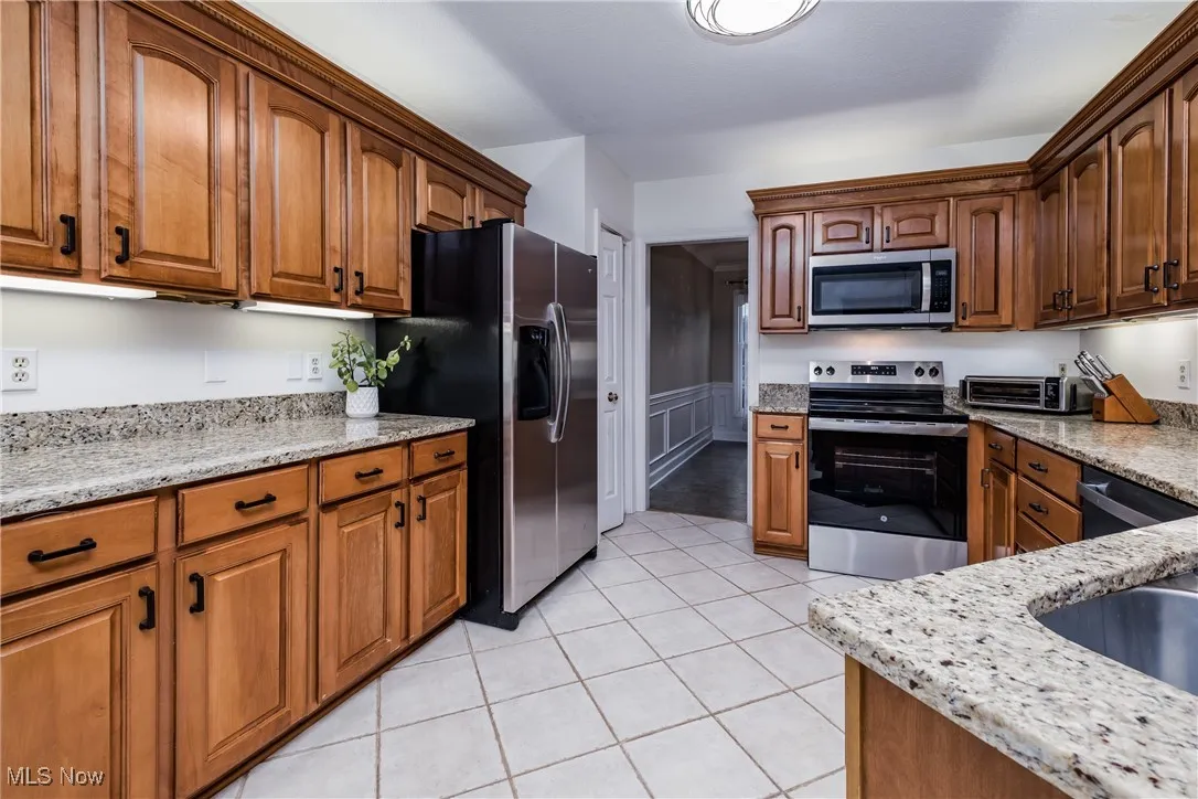 Kitchen with stainless steel appliances, light stone counters, light tile patterned floors, and brown cabinetry