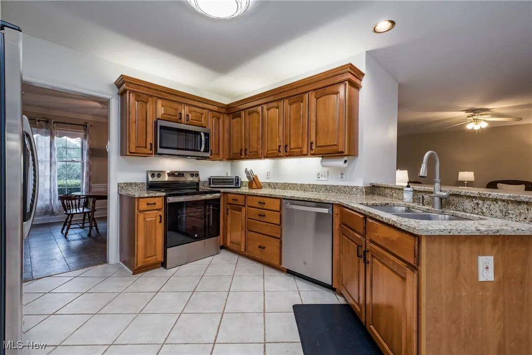 Kitchen featuring stainless steel appliances, light stone counters, brown cabinetry, ceiling fan, and light tile patterned floors