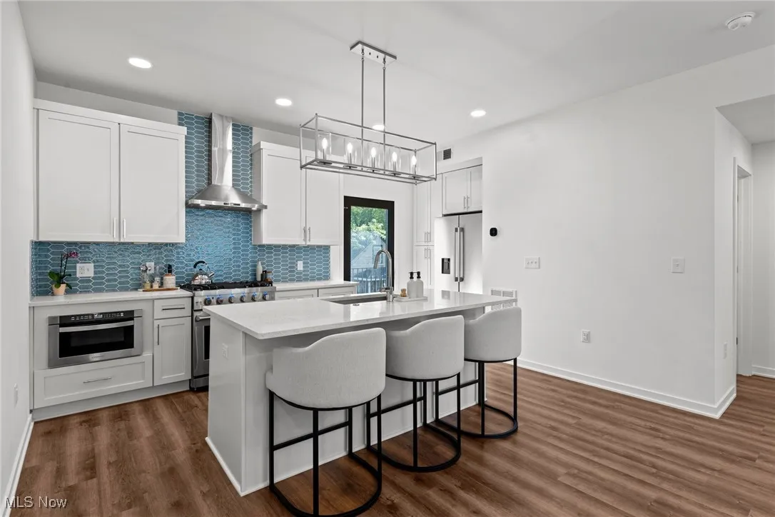 Kitchen featuring high quality appliances, wall chimney range hood, light countertops, a kitchen island with sink, and a kitchen bar