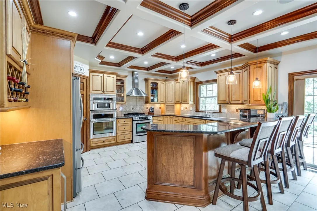 Kitchen with appliances with stainless steel finishes, wall chimney range hood, a peninsula, beam ceiling, and decorative backsplash