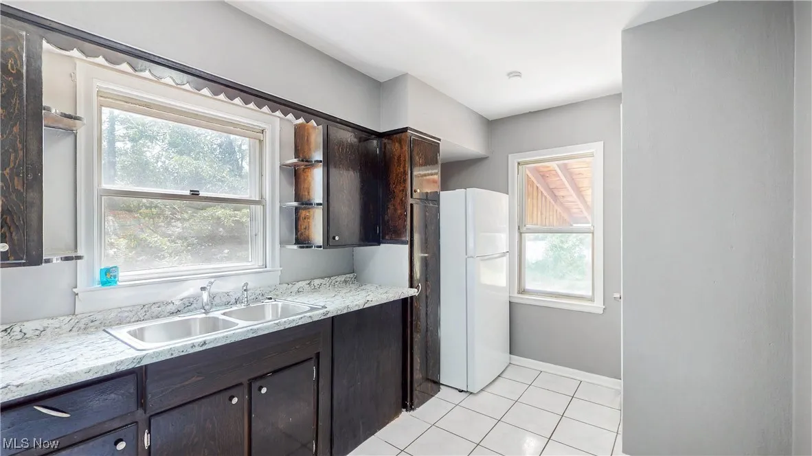 Kitchen with light tile patterned floors, freestanding refrigerator, and dark brown cabinets