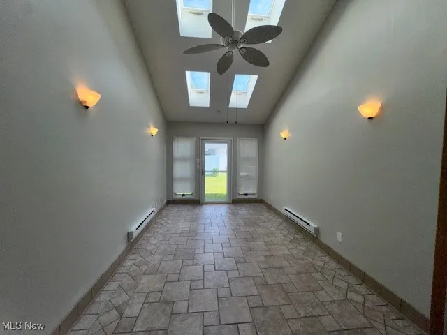 Unfurnished room featuring a skylight, high vaulted ceiling, baseboard heating, a ceiling fan, and stone finish flooring