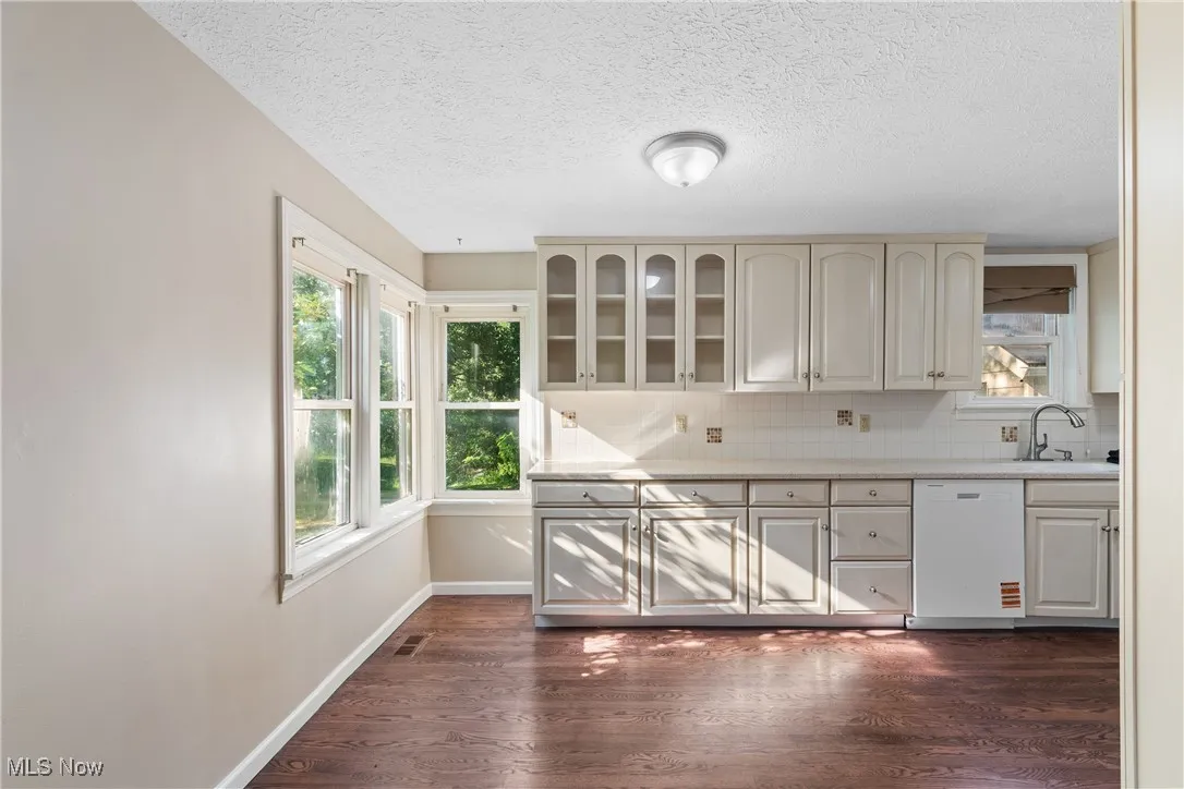 Kitchen featuring white dishwasher, light countertops, dark wood finished floors, decorative backsplash, and glass insert cabinets