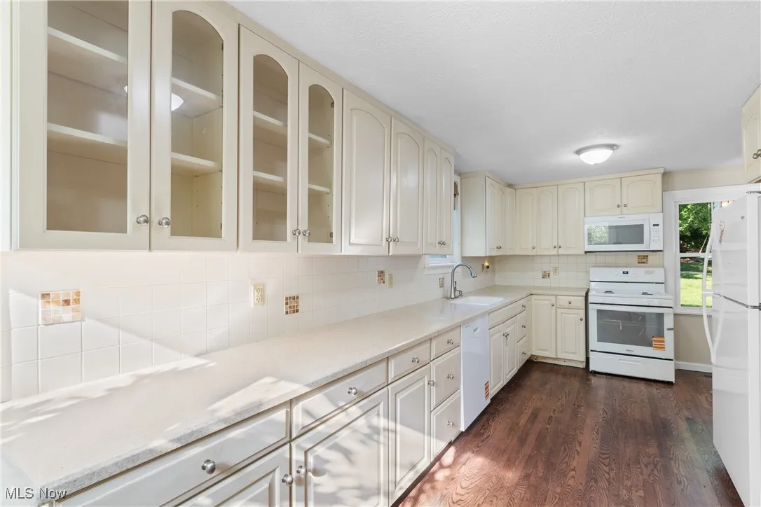 Kitchen featuring white appliances, dark wood finished floors, decorative backsplash, cream cabinetry, and glass insert cabinets