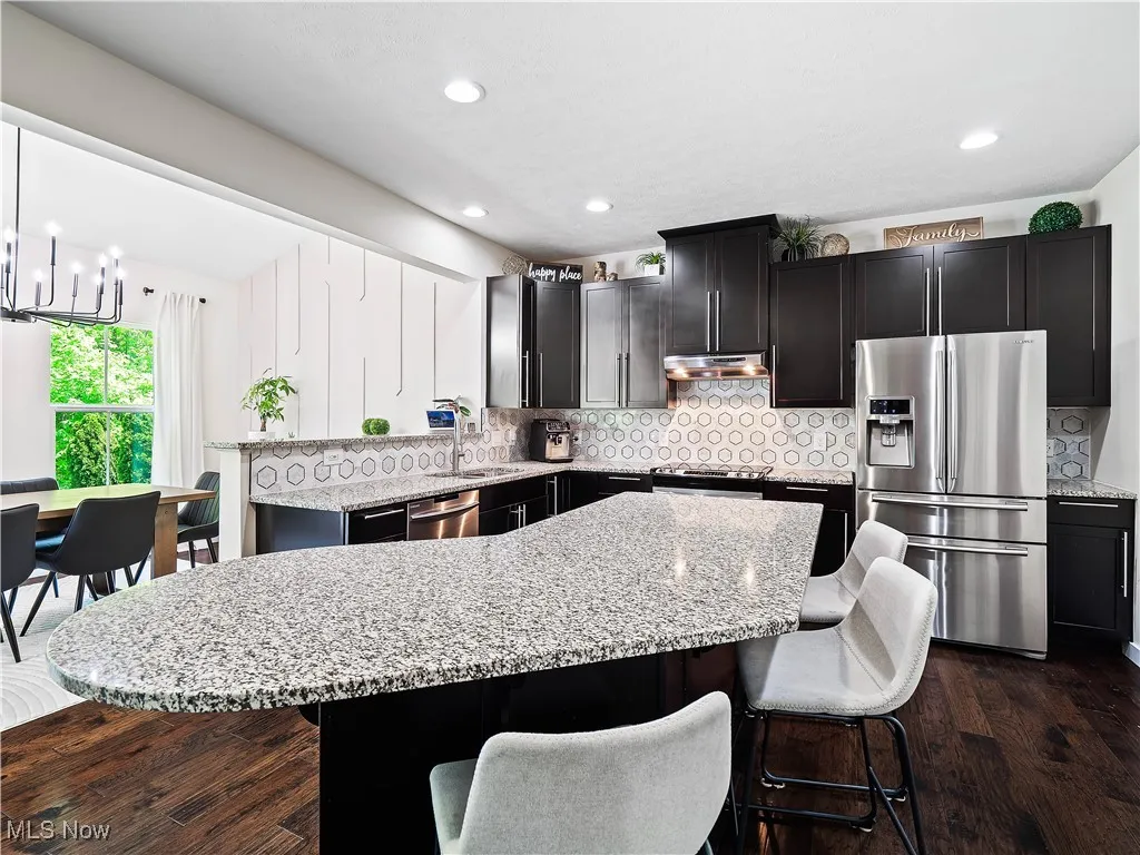 Kitchen with appliances with stainless steel finishes, a chandelier, a kitchen breakfast bar, light stone countertops, and under cabinet range hood