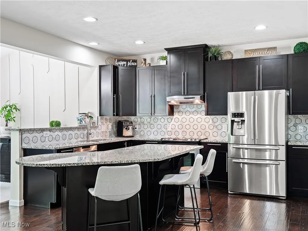 Kitchen featuring stainless steel fridge, under cabinet range hood, light stone counters, a kitchen bar, and a center island