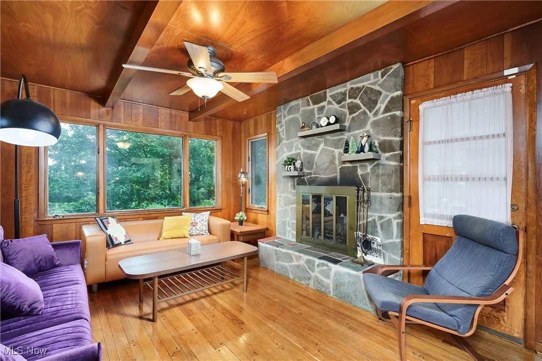 Living room with wooden walls, wood-type flooring, a wooden ceiling with exposed beams, a fireplace, and a ceiling fan
