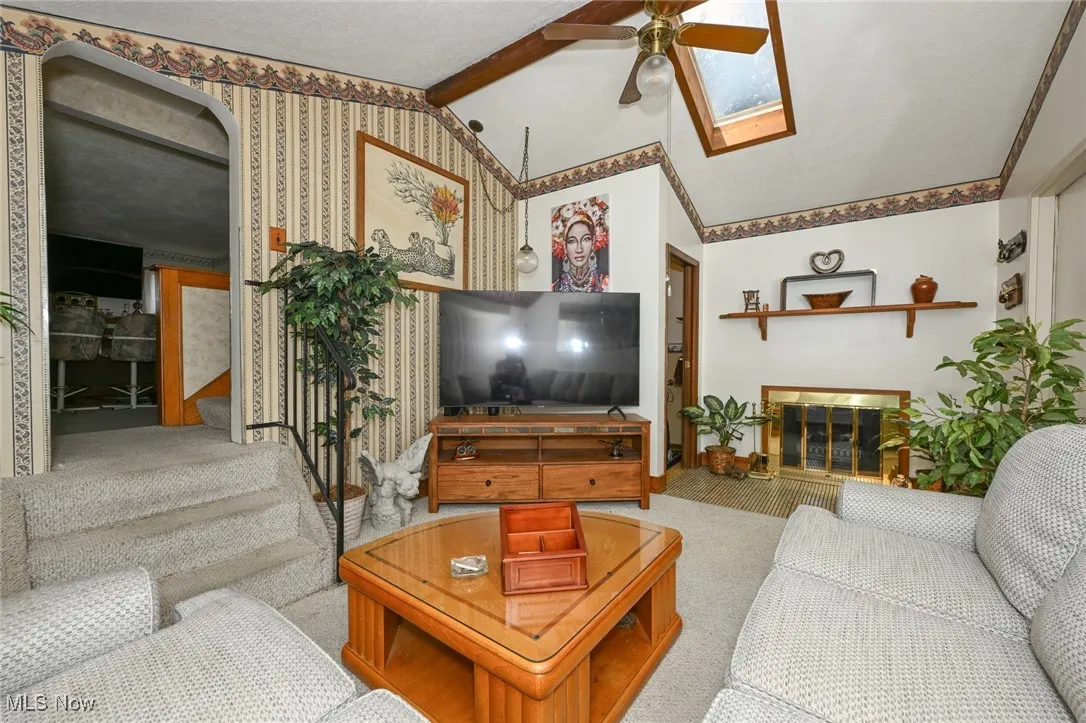 Living room with carpet flooring, a ceiling fan, stairs, a glass covered fireplace, and a skylight
