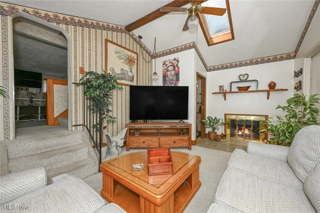 Carpeted living room featuring a ceiling fan, a glass covered fireplace, a skylight, and stairway