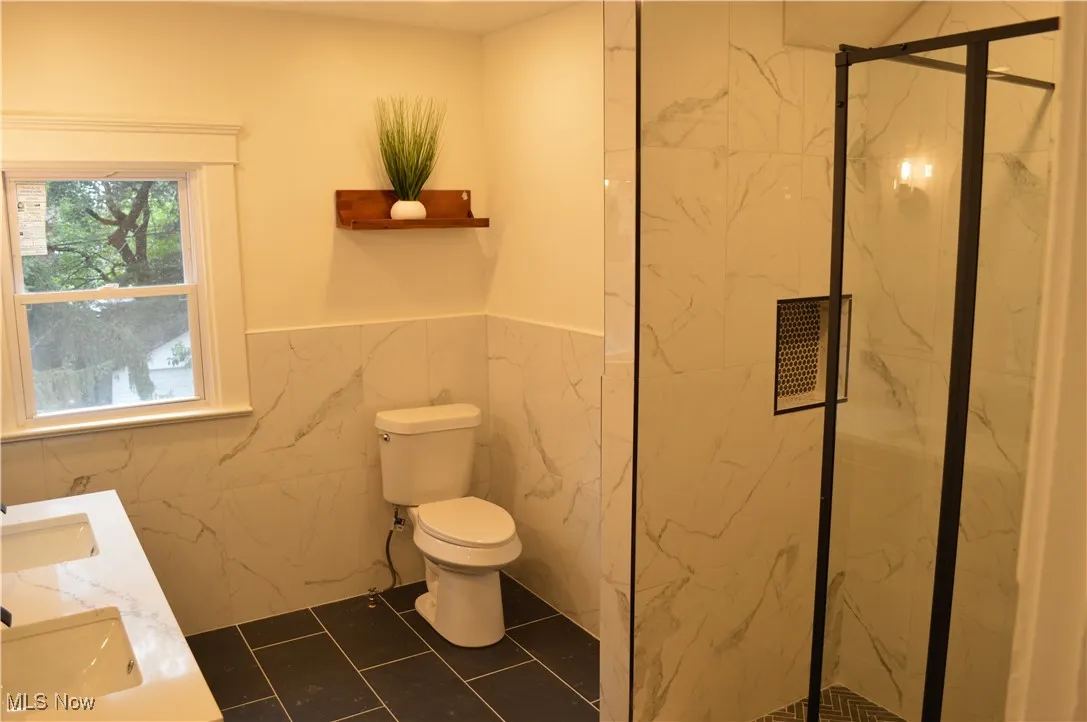 Bathroom with a marble finish shower, a wainscoted wall, tile walls, tile patterned floors, and double vanity
