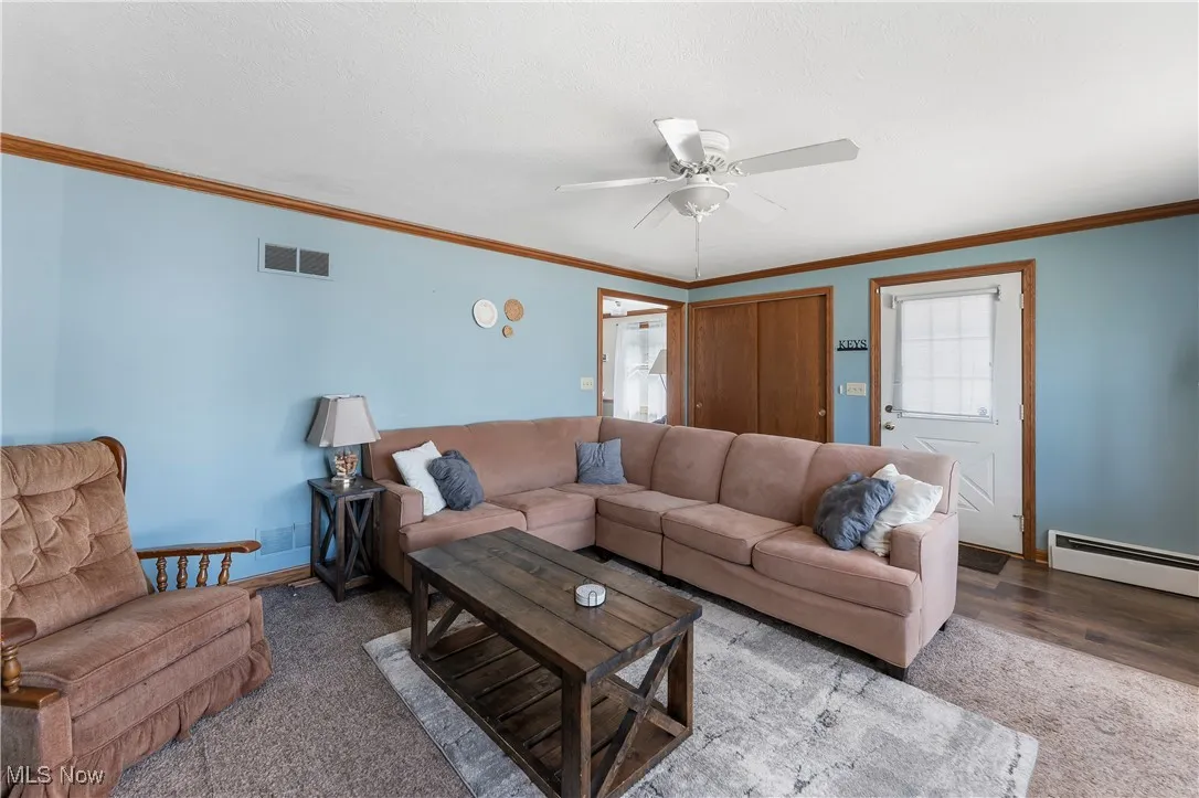 Living room featuring a baseboard heating unit, ceiling fan, carpet, and crown molding