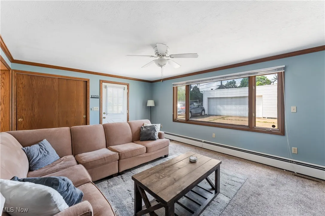 Living area featuring a baseboard heating unit, ceiling fan, light colored carpet, ornamental molding, and a textured ceiling
