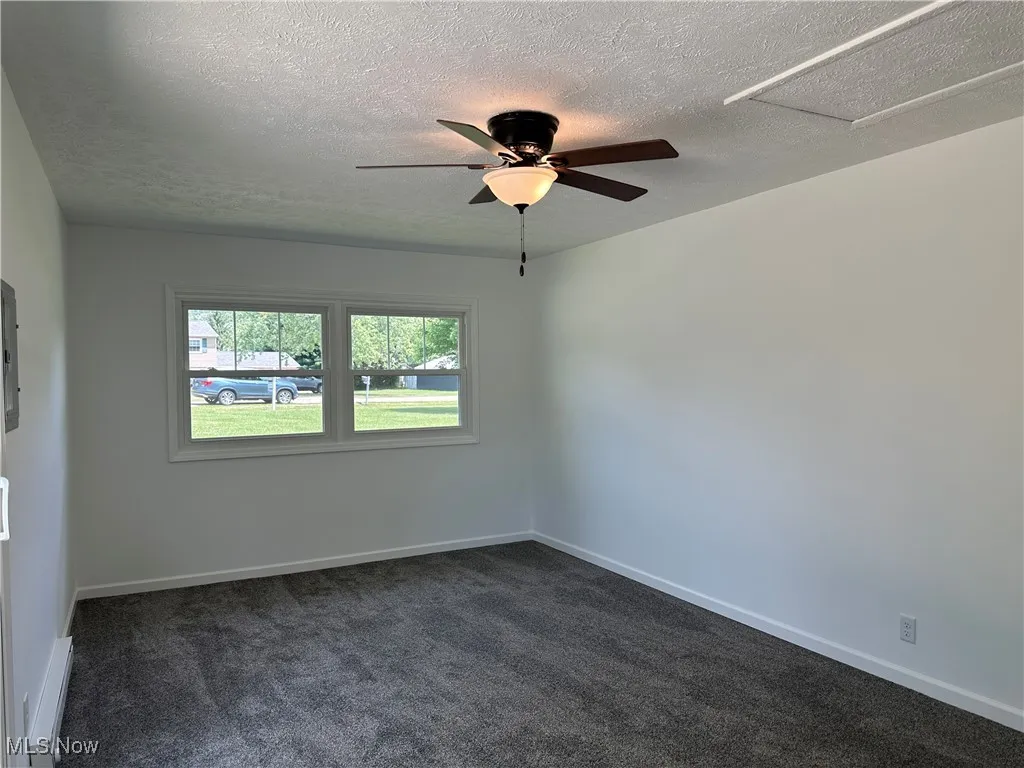 Empty room featuring a ceiling fan, a textured ceiling, dark carpet, a baseboard heating unit, and attic access
