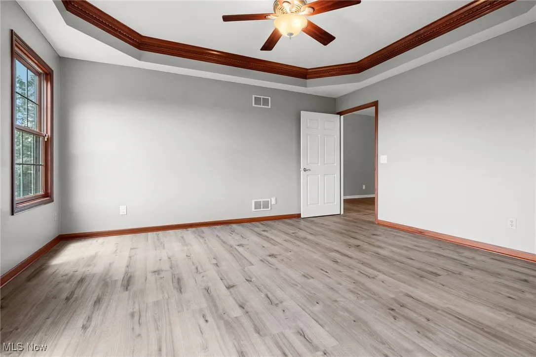 Empty room with a raised ceiling, ornamental molding, ceiling fan, and light wood-style flooring