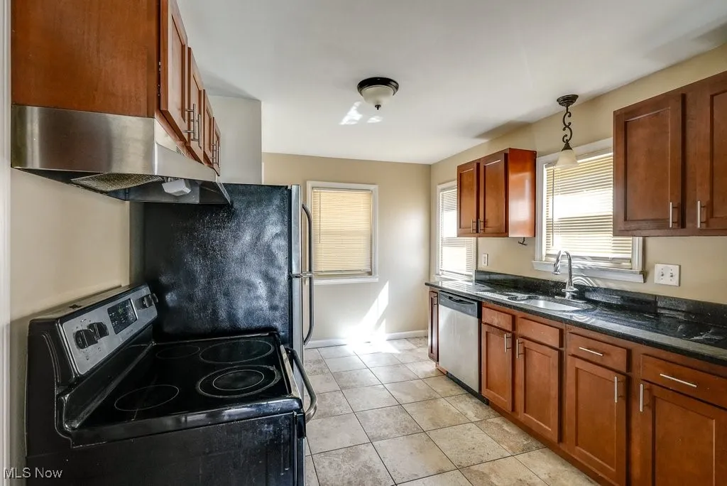 Kitchen featuring stainless steel appliances, under cabinet range hood, brown cabinets, light tile patterned floors, and decorative light fixtures