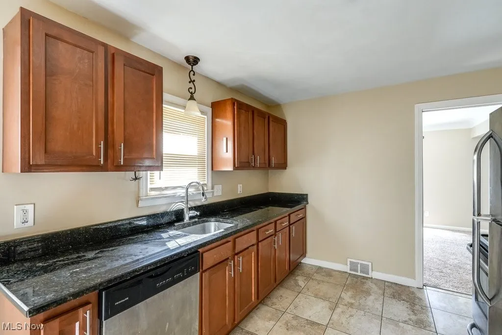 Kitchen featuring stainless steel dishwasher, brown cabinetry, dark stone counters, decorative light fixtures, and fridge