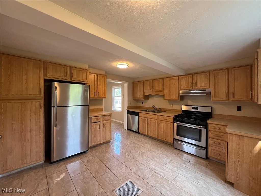 Kitchen with stainless steel appliances, under cabinet range hood, light countertops, a textured ceiling, and brown cabinets
