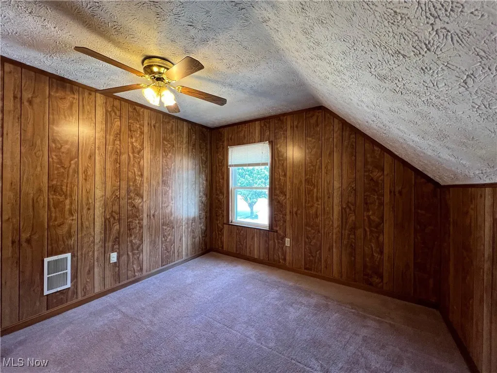 Additional living space featuring a textured ceiling, vaulted ceiling, ceiling fan, wooden walls, and carpet
