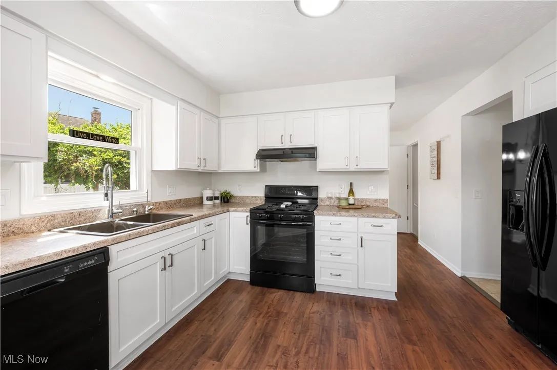 Kitchen with black appliances, under cabinet range hood, light countertops, dark wood-style flooring, and white cabinets