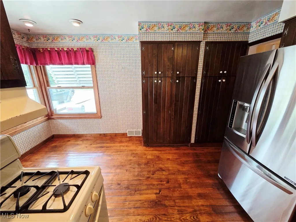 Kitchen with stainless steel fridge, white gas range, wallpapered walls, dark brown cabinetry, and dark wood-style flooring