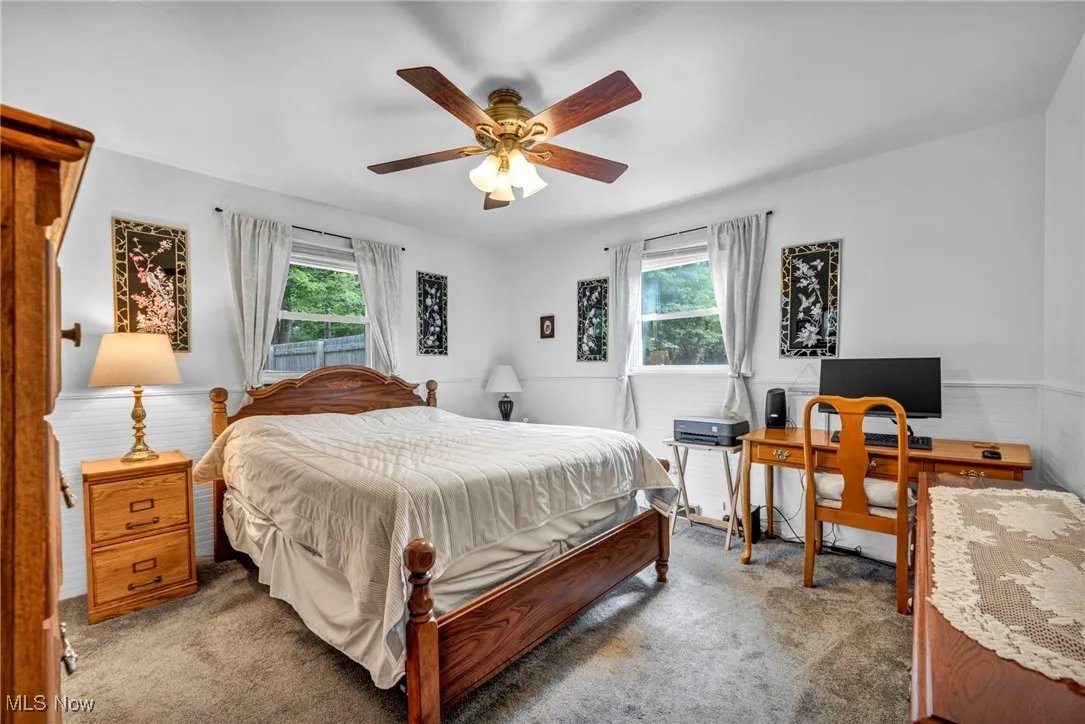 Carpeted bedroom with multiple windows, an office area, a ceiling fan, and wainscoting