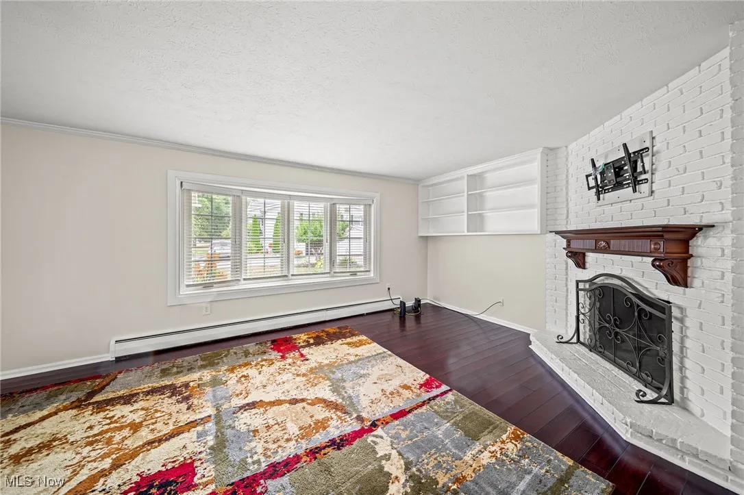 Living area featuring a baseboard heating unit, wood-type flooring, a textured ceiling, a brick fireplace, and crown molding