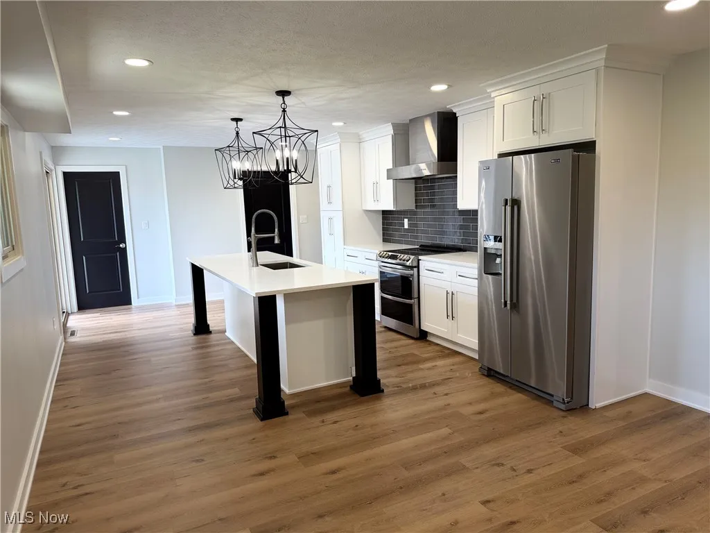 Kitchen with stainless steel appliances, wall chimney range hood, an island with sink, white cabinetry, and backsplash